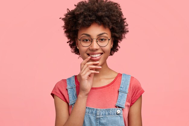 Alegre mujer rizada de piel oscura con una sonrisa agradable, vestida con una camiseta casual con un mono de mezclilla, aislada sobre una pared rosa.