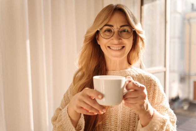 Alegre mujer rubia adulta de piel clara con gafas sonriendo a la cámara mientras bebe café aromático mientras está de pie junto a la ventana Relajación de ocio y concepto de estilo de vida