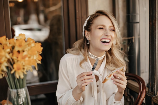 Alegre mujer rubia feliz en blusa blanca se ríe sinceramente sostiene cuchillo y paz de pan y descansa en el café de la calle