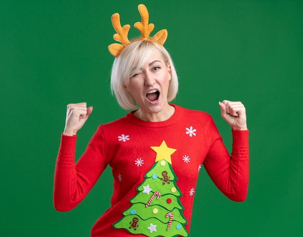 Alegre mujer rubia de mediana edad con diadema de cuernos de reno de Navidad y suéter de Navidad mirando a cámara guiñando un ojo haciendo gesto sí aislado sobre fondo verde