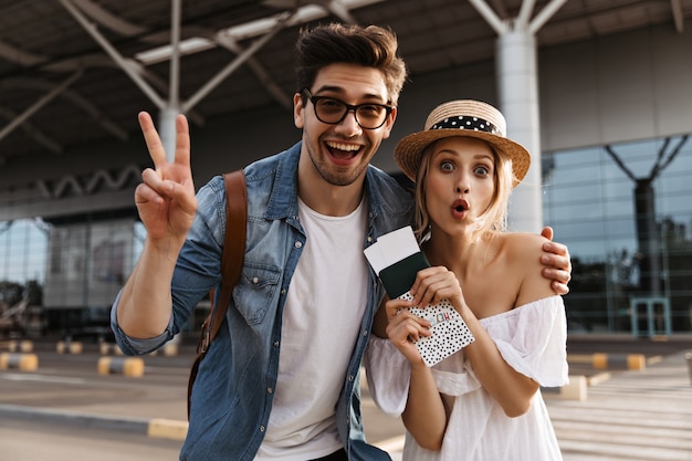 Alegre mujer rubia con sombrero hace una mueca, tiene pasaporte y boletos