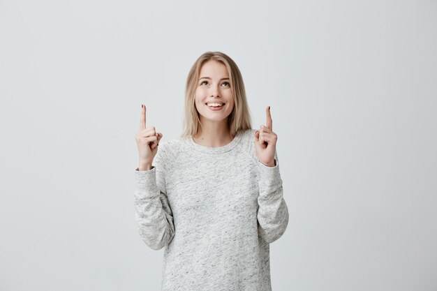 Alegre mujer rubia con suéter de manga larga mirando hacia arriba, señalando con el dedo el espacio de la copia por encima de su cabeza.