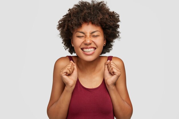 Alegre mujer satisfecha con piel oscura, celebra algo con los puños cerrados, tiene una amplia sonrisa