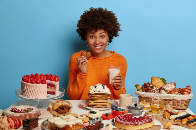 Alegre mujer sonriente con peinado afro rizado come sabrosos pasteles con leche, tiene buen humor para comer deliciosos postres, prueba deliciosas galletas recién horneadas