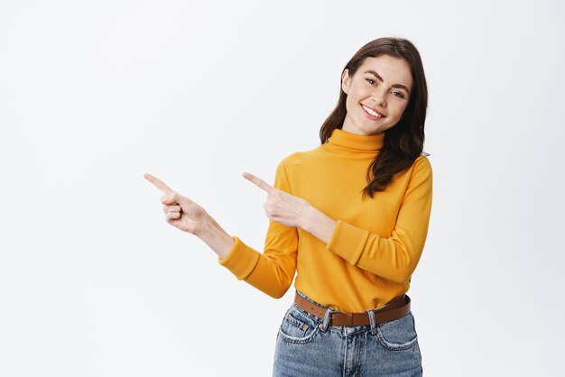 Alegre mujer sonriente señalando con el dedo el espacio de la copia y mirando a la cámara con una expresión agradable y amistosa de pie contra el fondo blanco Concepto de publicidad