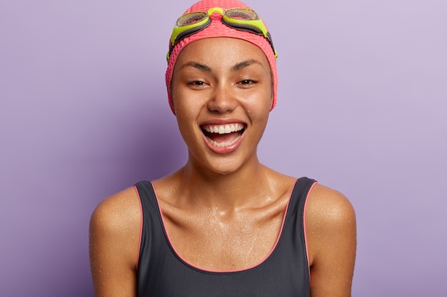 Alegre nadadora profesional deportista mojada después del entrenamiento en la piscina, viste traje de baño, sombrero y gafas, muestra dientes blancos, aislados sobre una pared púrpura. Horario de verano y concepto de descanso activo.