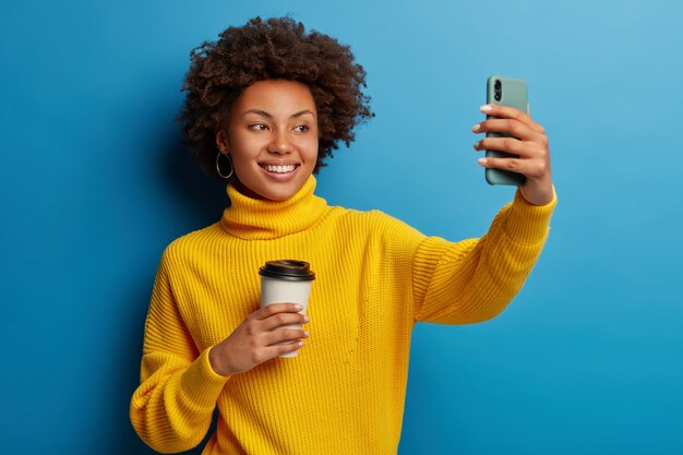 Alegre niña afro graba videos en línea, se toma una selfie en el teléfono móvil, extiende el brazo con un dispositivo moderno, se fotografía a sí misma, sostiene una taza de papel con café