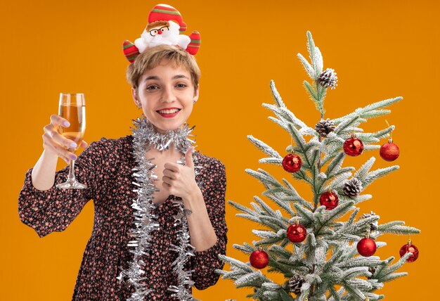 Alegre niña bonita con diadema de Papá Noel y guirnalda de oropel alrededor del cuello de pie cerca del árbol de Navidad decorado sosteniendo una copa de champán mostrando el pulgar hacia arriba aislado en la pared naranja