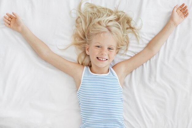 Alegre niña con cabello claro, acostada en una cama cómoda sobre sábanas blancas, estirando después de dormir por la noche, mirando con expresión encantadora. Niño pequeño pecoso descansando en la cama