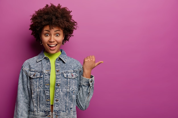 Alegre niña de cabello rizado señala con el pulgar hacia la derecha, muestra el área del espacio de la copia, se ríe positivamente, usa una chaqueta de mezclilla, aislada sobre una pared púrpura, demuestra un bonito anuncio contra la pared púrpura