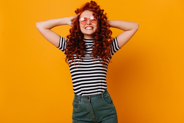 Alegre niña de jengibre sonríe sinceramente, posando en jeans, top y gafas de sol en el espacio naranja.