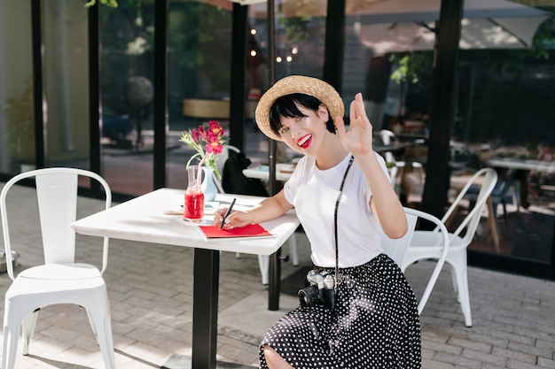 Alegre niña morena escribe en el cuaderno y agitando la mano con una sonrisa, descansando en el café