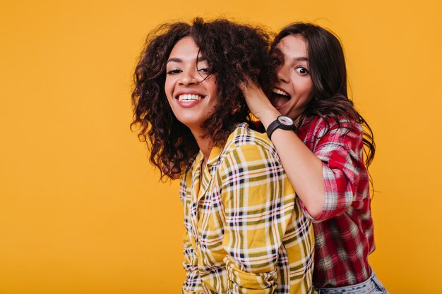 Alegre niña morena de ojos marrones se divierte y juega con el cabello de su mejor amiga. Mulato está sonriendo y mirando.