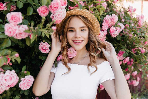 Alegre niña de ojos azules posando en la naturaleza y sonriendo. Impresionante modelo de mujer en sombrero de paja de moda relajante sabor a flores.