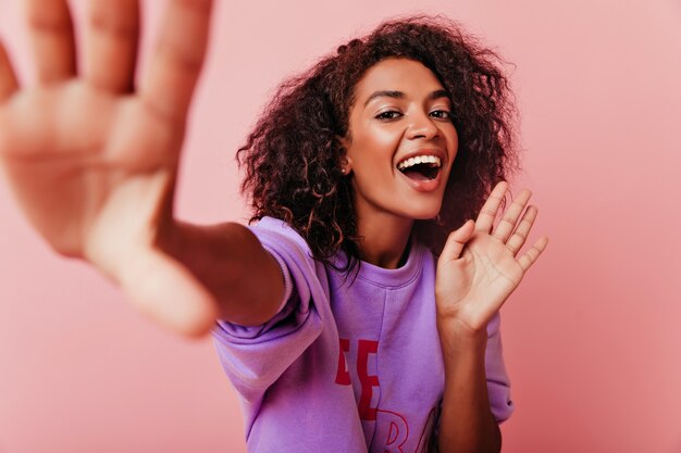 Alegre niña de ojos oscuros sonriendo mientras hace selfie. Increíble dama africana morena jugando en el estudio.