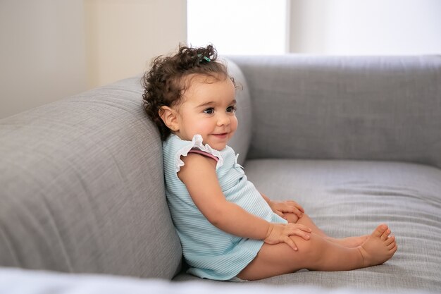 Alegre niña de pelo rizado oscuro vistiendo un paño azul pálido, sentada en un sofá gris en casa, mirando a otro lado y sonriendo. Niño en casa y concepto de infancia.