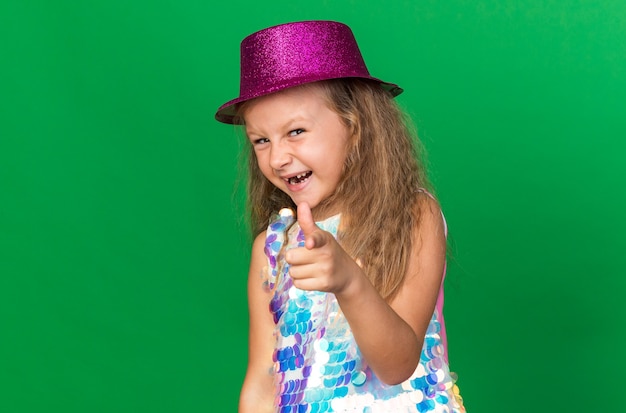 Alegre niña rubia con gorro de fiesta púrpura apuntando aislado en la pared verde con espacio de copia