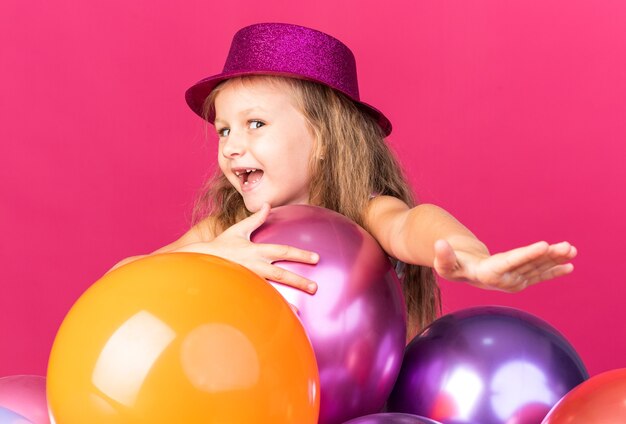 Alegre niña rubia con gorro de fiesta púrpura de pie con globos de helio aislado en la pared rosa con espacio de copia