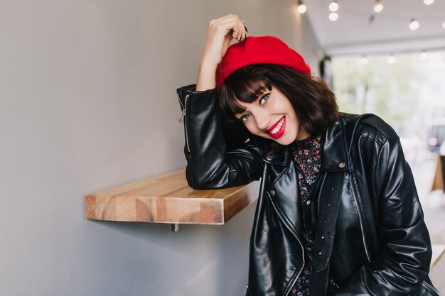 Alegre niña sonriente con cabello castaño oscuro y ropa de estilo rock posando junto al estante de madera en la habitación luminosa. Retrato de adorable joven morena vistiendo boina francesa y chaqueta de cuero negro