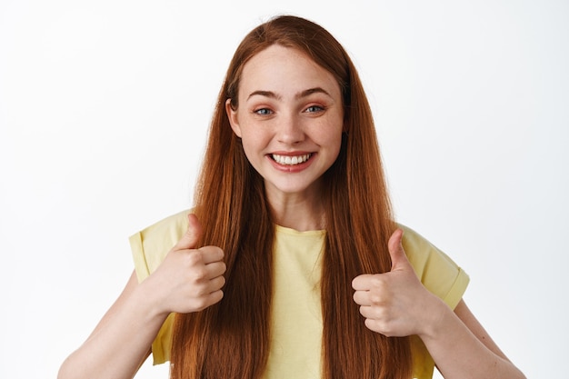 Alegre niña sonriente con el pelo rojo, mostrando los pulgares hacia arriba en señal de aprobación, decir que sí, luciendo satisfecha, alabanza y elección de cumplidos, de pie en aislado.