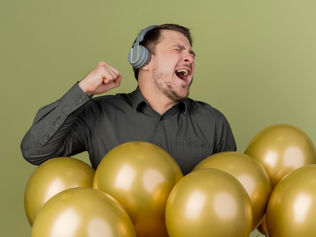 Alegre con los ojos cerrados chico joven partido con camisa negra de pie entre globos escuchar música de auriculares aislados en verde oliva