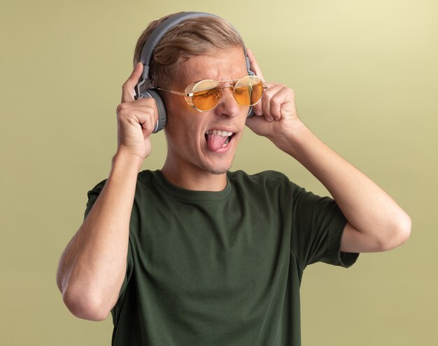 Alegre con los ojos cerrados joven guapo con camisa verde con gafas y auriculares mostrando la lengua aislada en la pared verde oliva