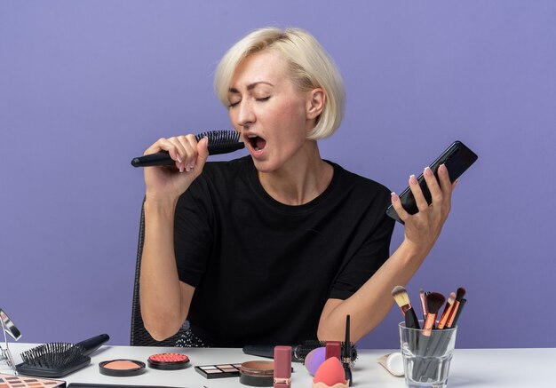 Alegre con los ojos cerrados joven hermosa se sienta a la mesa con herramientas de maquillaje sosteniendo el teléfono con peine y cantando aislado en la pared azul