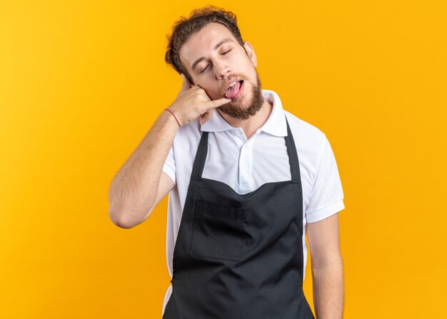 Alegre con los ojos cerrados joven peluquero vistiendo uniforme mostrando lengua y gesto de llamada telefónica aislado en la pared amarilla