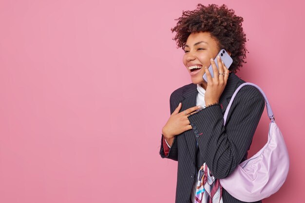 Alegre y optimista mujer de pelo rizado en ropa formal se ríe felizmente tiene una conversación telefónica lleva una bolsa sostiene un teléfono inteligente cerca de la oreja aislada sobre un fondo rosa con un espacio vacío para su texto