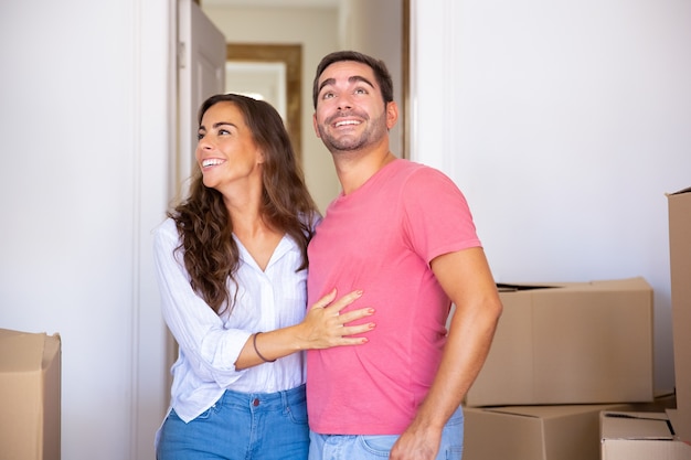 Foto gratuita alegre pareja emocionada mudarse a la nueva casa, de pie entre una caja de cartón, abrazándose y mirando a su alrededor