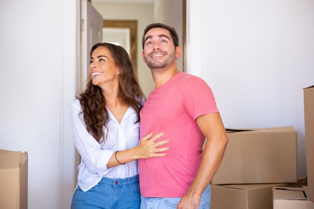 Alegre pareja emocionada mudarse a la nueva casa, de pie entre una caja de cartón, abrazándose y mirando a su alrededor