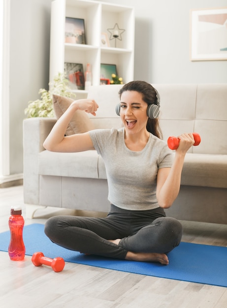 Alegre parpadeó haciendo un gesto fuerte joven con auriculares haciendo ejercicio con mancuernas en la estera de yoga en el sofá delantero en la sala de estar