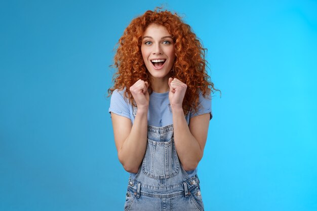 Alegre pelirroja afortunada optimista mujer atractiva de pelo rizado animando aprieta los puños con alegría sonriendo ampliamente animando viendo el juego de apoyo animar a mantener el fondo azul motivado.