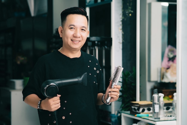 Alegre peluquero masculino asiático posando con secador de pelo y cepillo en el salón