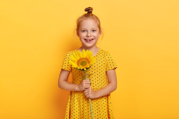 Alegre pequeña niña pelirroja posando con girasol en vestido amarillo