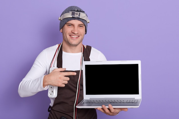 alegre radioman positivo sosteniendo portátil con pantalla en blanco, haciendo gesto, mostrando con el dedo índice, con doble adaptador y varios cables en el cuello, de pie con el equipo necesario.