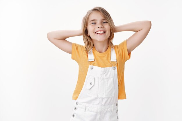 Alegre, relajada, bonita, rubia, niña, relajarse, recostarse y disfrutar de las vacaciones de verano, días libres de escuela los fines de semana, descansar en las vacaciones, tomarse de las manos detrás de la cabeza, pose perezosa sonriendo felizmente, riendo