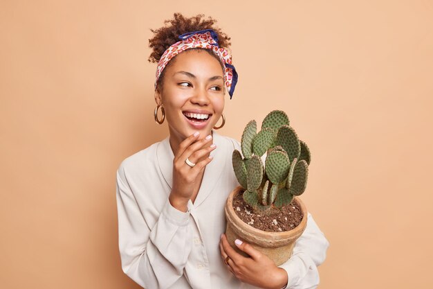 Alegre y sincera mujer afroamericana lleva maceta de planta de interior le gusta el cactus