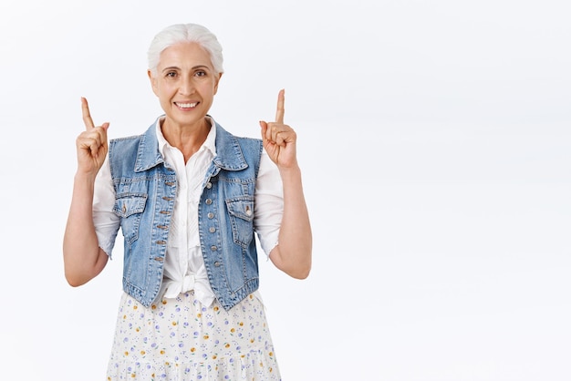 Foto gratuita alegre sonriente anciana caucásica en chaleco de mezclilla sobre vestido sonriendo feliz señalando con el dedo hacia arriba recomendar producto sugerir enlace útil mostrando orgullosamente promo fondo blanco de pie