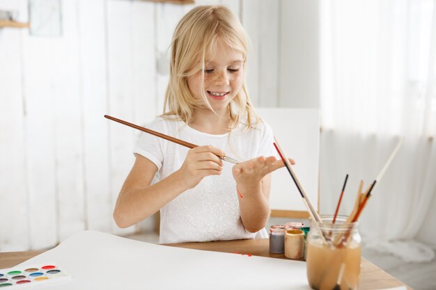 Alegre, sonriente y feliz niña rubia en camiseta blanca dibujando algo en su palma con un pincel.