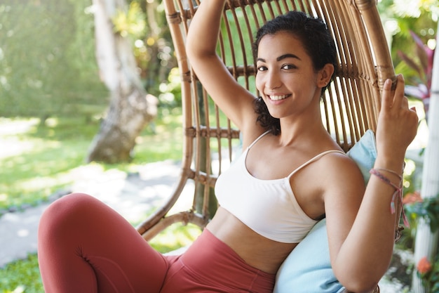 Alegre tierna mujer feliz con cabello oscuro y rizado disfrutando de unas vacaciones sentado en una silla de ratán