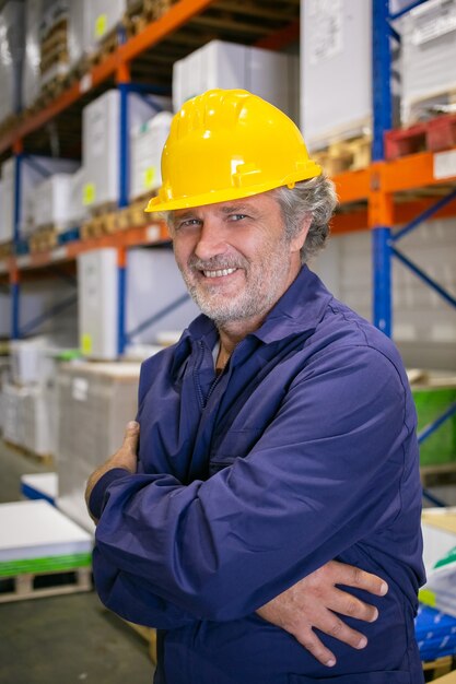 Alegre trabajador logístico de pelo gris en casco y uniforme de pie en los estantes del almacén con los brazos cruzados, mirando a cámara y sonriendo. Disparo vertical. Concepto de retrato de trabajo y cuello azul