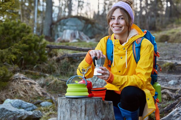 Alegre turista mantenga la cafetera, tiene un picnic en el muñón, usa una bufanda en la cabeza, un impermeable amarillo