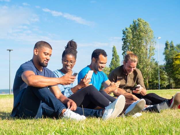 Alegres amigos multiétnicos que usan teléfonos inteligentes en el parque