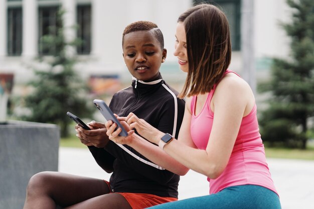 Alegres amigos sonrientes en ropa deportiva sentados en un banco en la ciudad discutiendo mientras usan un teléfono inteligente mirando a la pantalla Mujeres multiétnicas que tienen un descanso de entrenamiento físico