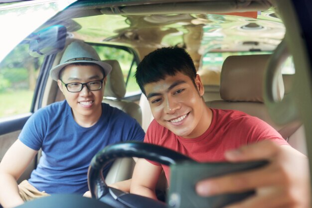 Alegres amigos varones asiáticos sentados juntos en el coche y tomando selfie