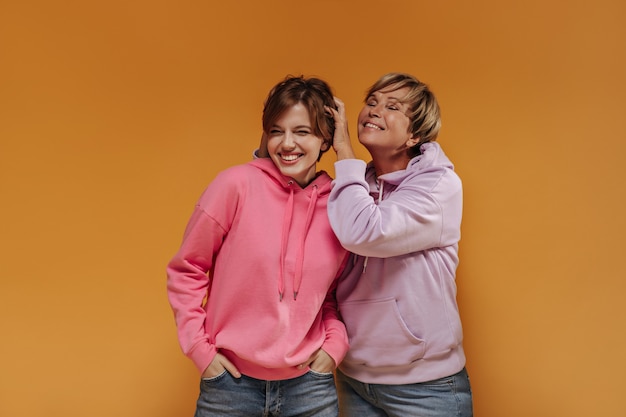 Alegres dos mujeres con pelo corto en amplias sudaderas con capucha elegantes y pantalones vaqueros frescos sonriendo y divirtiéndose sobre fondo naranja aislado.