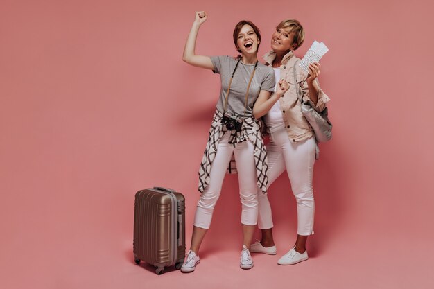 Alegres dos mujeres con el pelo corto en pantalones blancos delgados y zapatillas de deporte sonriendo y posando con maleta, cámara, boletos y bolso sobre fondo aislado.