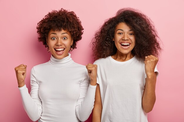 Alegres felices dos aturdidores con cabello afro, mantienen los brazos en alto, aprietan los puños con éxito