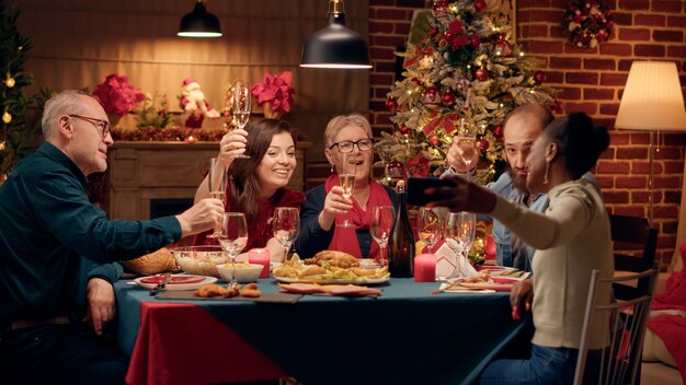 Alegres miembros multirraciales de la familia tomando fotos de selfie con un dispositivo de teléfono inteligente mientras se sientan en la mesa de la cena de Navidad. Diversas personas festivas celebrando las vacaciones de invierno juntas en casa.